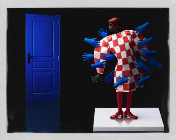 Model in a red and white checkered coat surrounded by reaching blue gloved hands next to a blue door against a black backdrop.