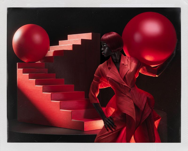 A Black model with a red bob haircut poses in a red pleated suit holding a large red sphere beside a red staircase set.