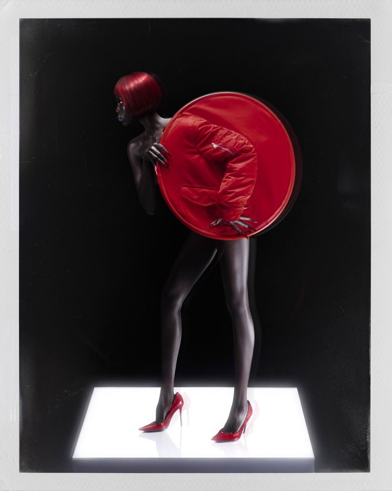 Model with red hair in a sculptural red circular garment and red stiletto heels stands on a glowing white platform against a black studio.