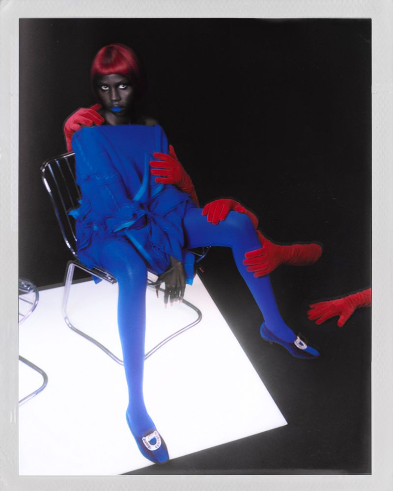 Model with red hair wearing a vibrant blue dress and tights seated on a chair, surrounded by disembodied red gloved hands.