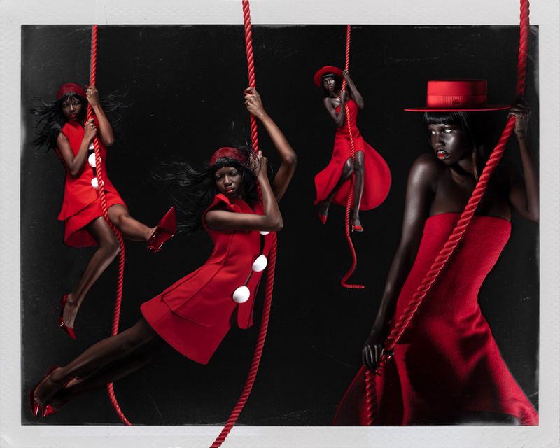 Black female models in red dresses and hats suspended from thick red ropes against a deep black studio backdrop.