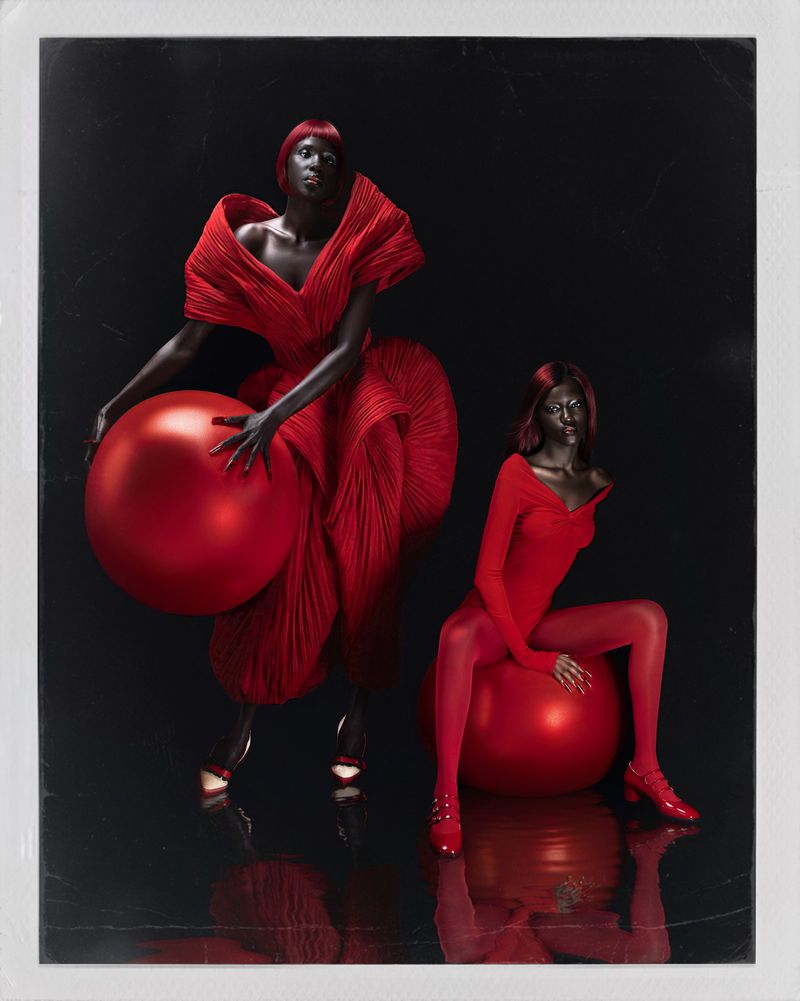 Two black female models in vibrant red sculptural outfits posing with large red spheres against a dark reflective studio background.