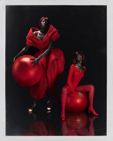Two black female models in vibrant red sculptural outfits posing with large red spheres against a dark reflective studio background.