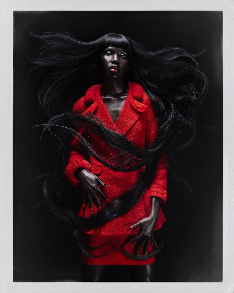 Fashion model with long windswept black hair wearing a structured red tweed suit against a solid black background.