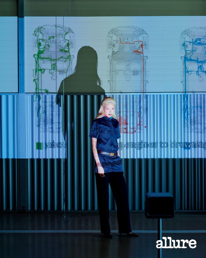 Model Minji Lee in a short-sleeved navy shirt and dark trousers posing before projected technical diagrams for Allure Korea.