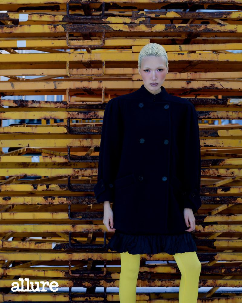 Model Minji Lee in an oversized black coat and yellow tights posing before stacked yellow metal scaffolding.