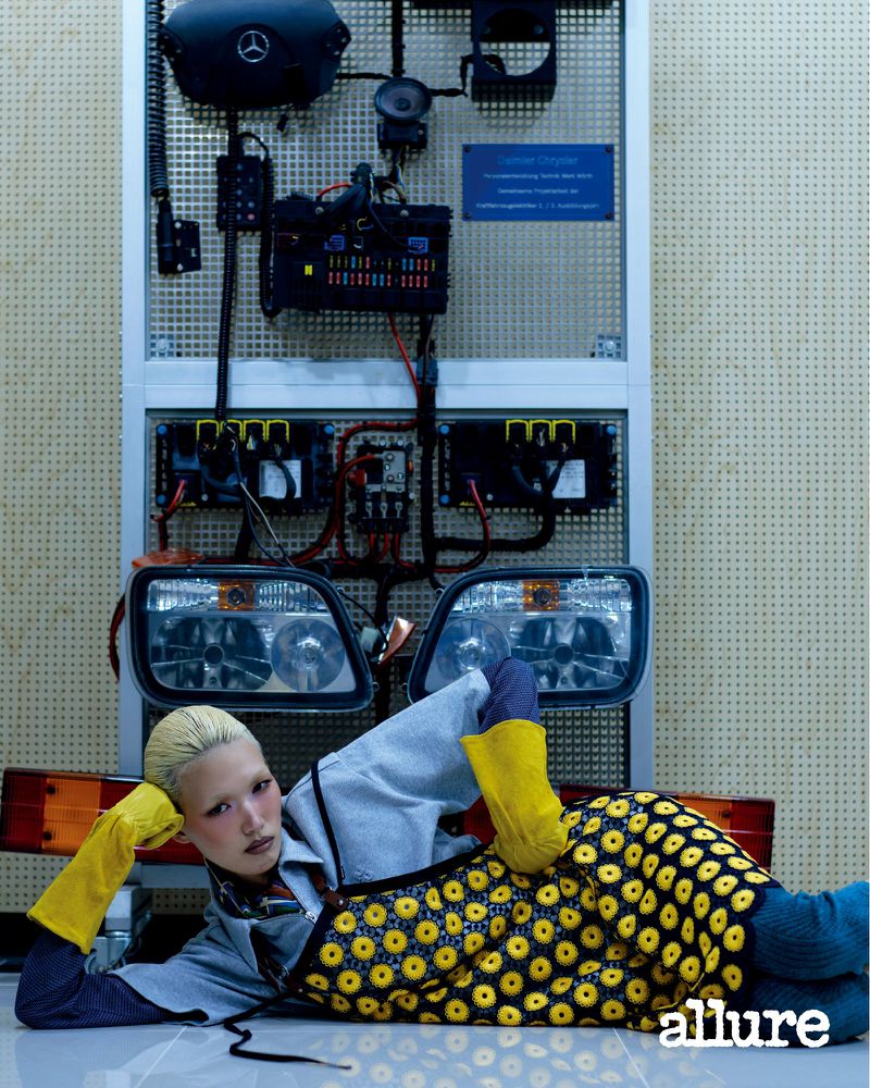 Model Minji Lee lying down wearing yellow rubber gloves and a patterned apron against an automotive electrical display board.
