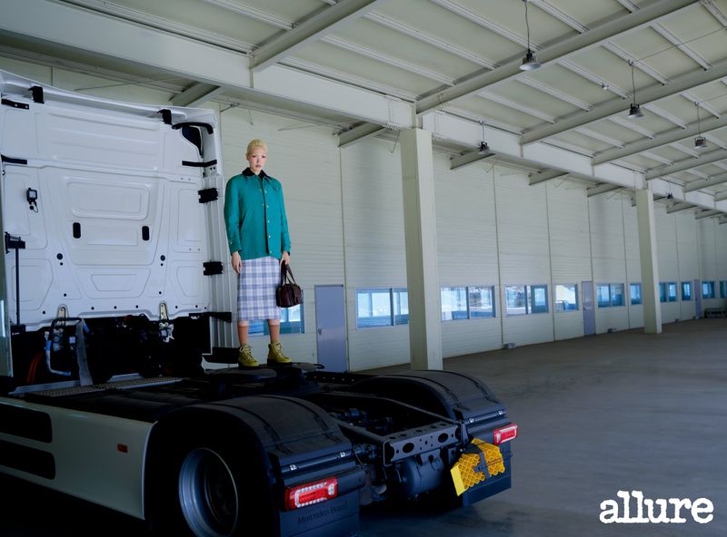 Model Minji Lee poses atop a white semi-truck chassis inside a spacious warehouse, wearing a green jacket and plaid skirt.