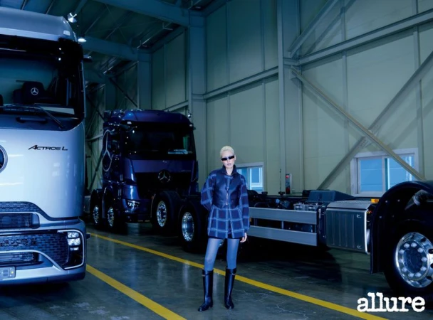 Minji Lee poses in a plaid jacket, leather vest, and tall boots inside a garage with large trucks for Allure Korea.
