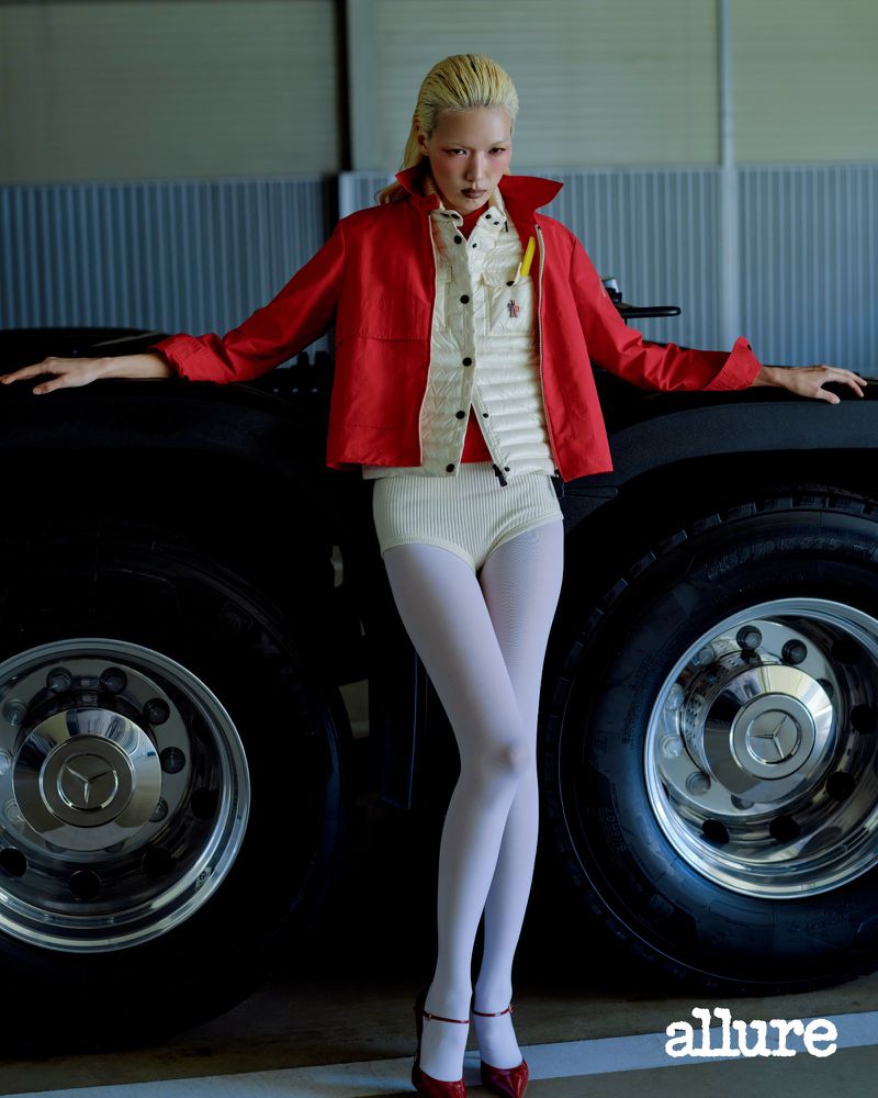 Model Minji Lee wearing a red jacket, cream quilted vest, white knit shorts, and white tights leaning on large truck tires.