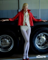 Model Minji Lee wearing a red jacket, cream quilted vest, white knit shorts, and white tights leaning on large truck tires.