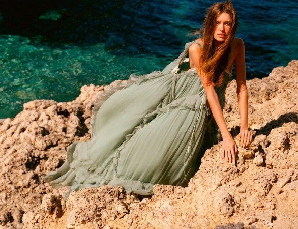 Model posing on a sunlit rocky cliff above clear turquoise water wearing a voluminous sage green ruffled dress.