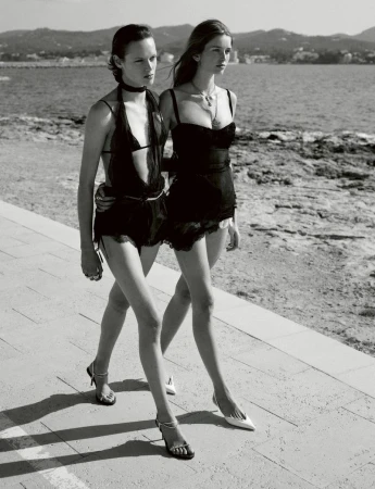 Two female models in black lace outfits walking together along a sunny coastal promenade in black and white.