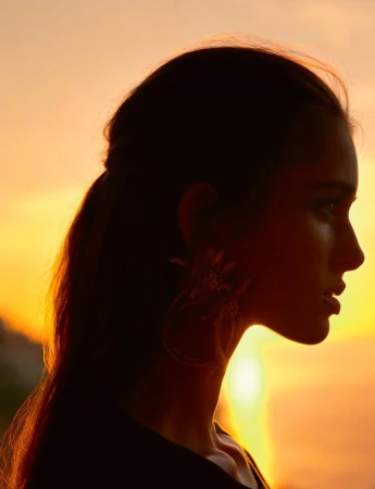 Profile silhouette of a woman wearing large statement earrings against a glowing golden orange sunset sky.