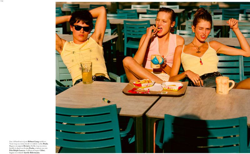 Three models seated at an outdoor terrace table with novelty mugs and snacks in direct sunlight for Harper's Bazaar France.