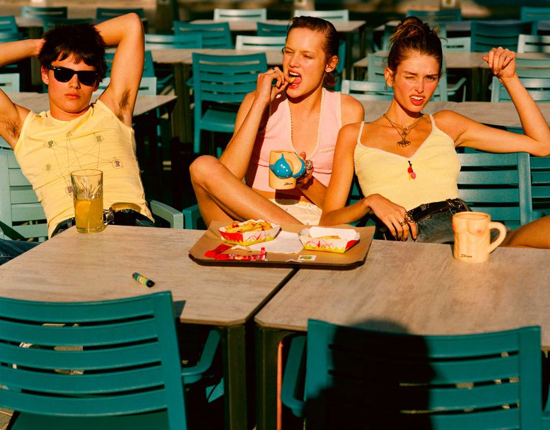 Three models wearing summer tank tops seated at an outdoor cafe table with novelty mugs and fast food snacks in bright sunlight.