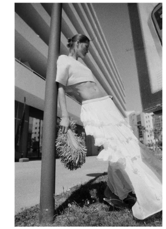 Model Bonnie Panasenko wearing a white crop top and tiered ruffle skirt leaning against a pole outdoors in black and white.