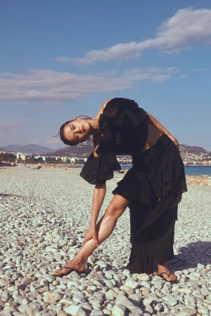 Model Bonnie Panasenko poses on a pebble beach wearing a black ruffled outfit and sandals under a blue sky.