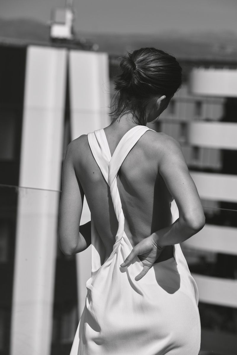 Monochrome rear view of a model in a white backless twisted-strap dress outdoors against modern architectural buildings.