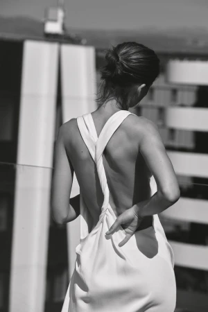 Monochrome rear view of a model in a white backless twisted-strap dress outdoors against modern architectural buildings.