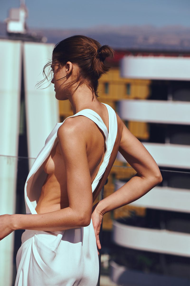 Model Bonnie Panasenko viewed from behind wearing a backless draped white top on a sunny outdoor rooftop.