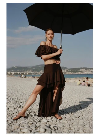 Model Bonnie Panasenko posing on a pebble beach holding a black umbrella while wearing a dark brown ruffled crop top and tiered skirt.