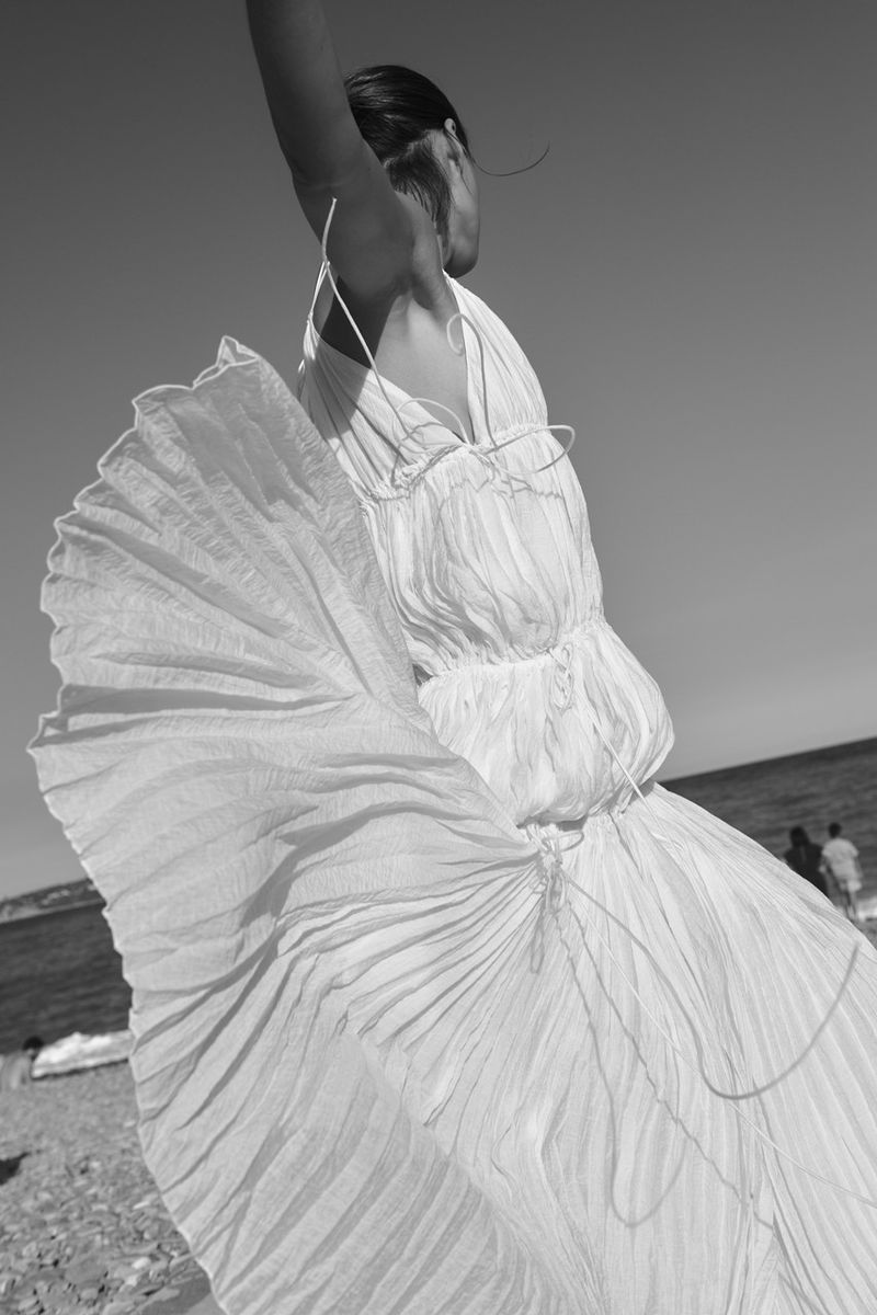 Black and white photo of a woman in a voluminous pleated white dress billowing in the wind on a rocky shoreline.