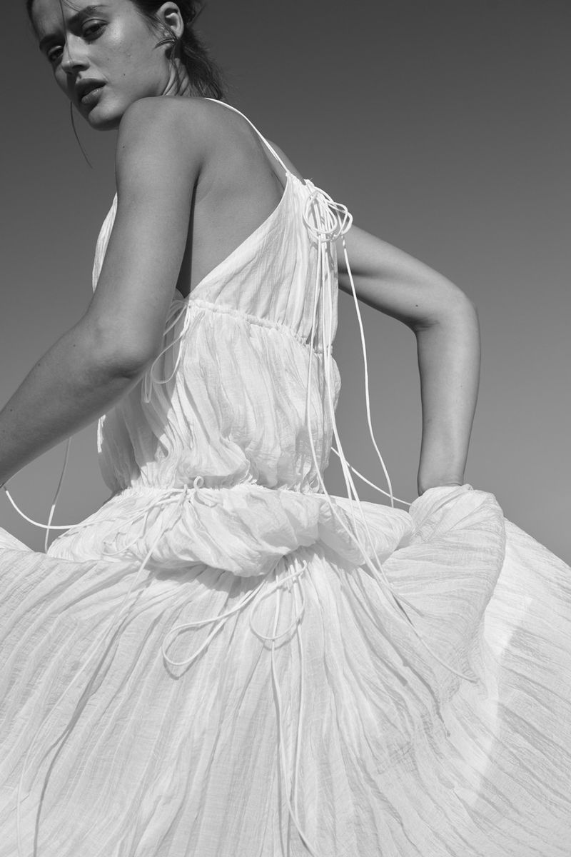 Model in a white pleated dress looking back outdoors against a clear sky for H and M Du matin au soir campaign.