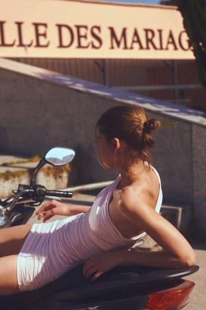 Model in a pink minidress resting on a scooter outdoors in front of a building exterior with text signage