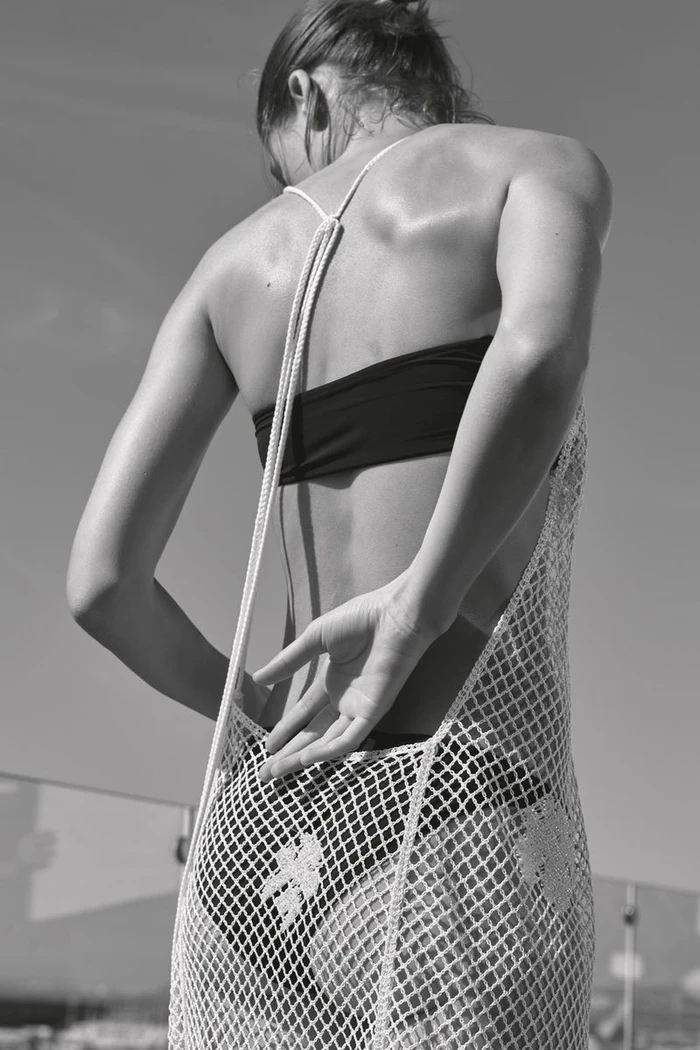 Black and white shot of model Bonnie Panasenko from behind wearing a black bikini and white crochet open-back dress.