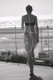 Model seen from behind wearing a black bikini and sheer white mesh cover-up walking on a sunlit wooden deck overlooking the ocean.