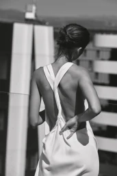 Monochrome rear view of a model in a white backless twisted-strap dress outdoors against modern architectural buildings.