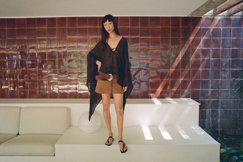 Model Diane Chiu in a sheer black tunic, brown suede mini skirt, and wide belt posing against an iridescent tiled wall.