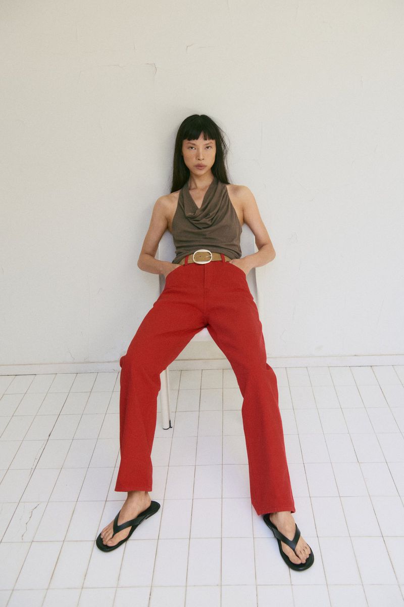 Model Diane Chiu seated in red trousers and a taupe cowl-neck top against a white studio wall with tiled flooring.