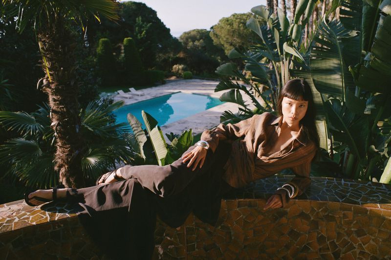 Model reclining against a mosaic ledge amidst lush tropical foliage with a swimming pool visible in the background.