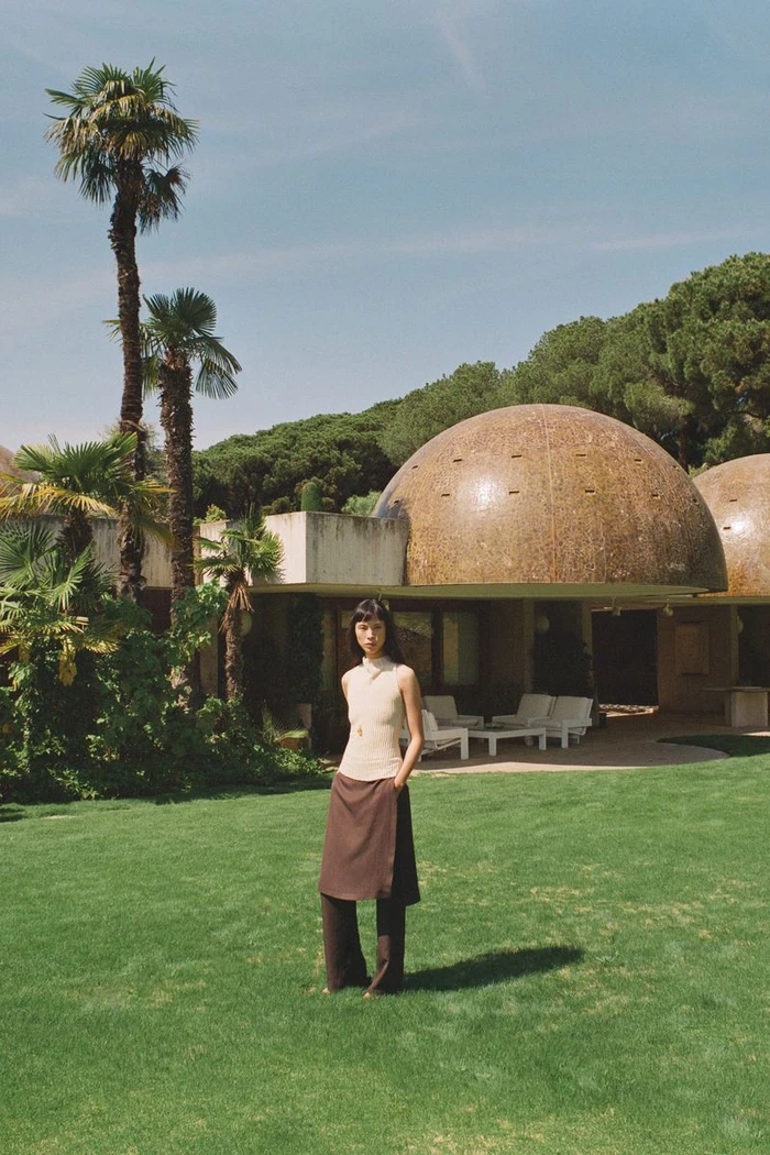 Model Diane Chiu standing on a green lawn wearing a sleeveless cream top and brown layered pants in front of a domed modernist house.