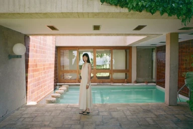 Diane Chiu in a cream sleeveless dress standing poolside on a stone patio outside a modernist villa.