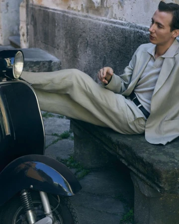 A man in a relaxed beige suit reclined on a stone bench with feet resting on a vintage black scooter.