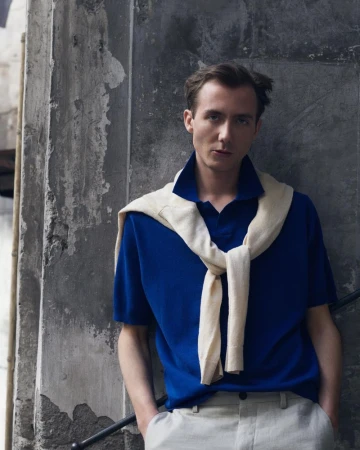 Model Paul Hameline wearing a vibrant blue polo shirt and a cream sweater draped over his shoulders against a weathered wall.