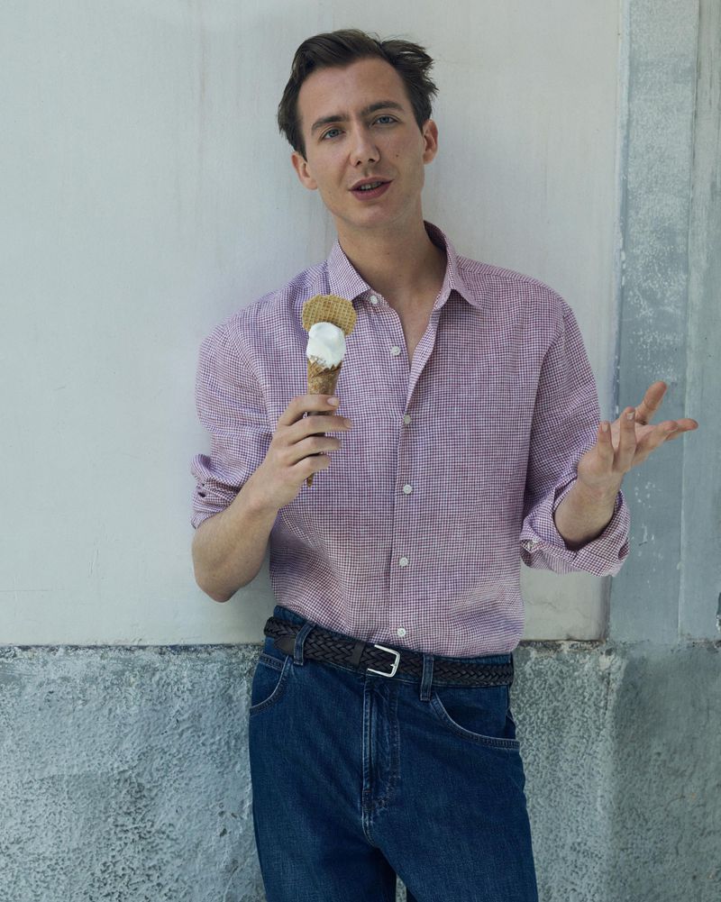 Young adult poses outdoors with an ice cream cone, wearing a button-down shirt and jeans, against a neutral textured wall backdrop.
