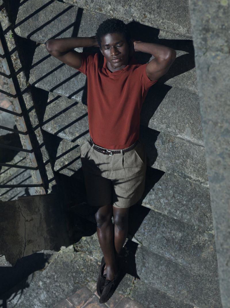 Model Mohammed Banze in a rust polo shirt and beige shorts leaning back on stone stairs under direct sunlight.