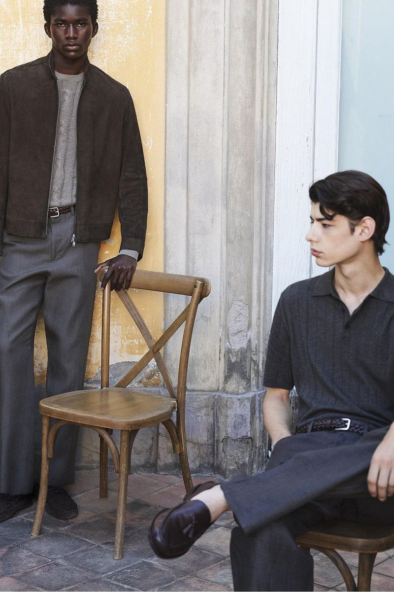Two male models in tailored menswear and knitwear posing with wooden chairs outdoors against a stone building wall.