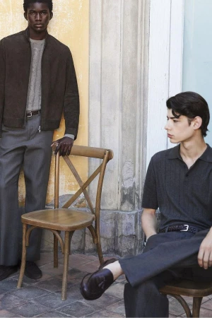 Two male models in tailored menswear and knitwear posing with wooden chairs outdoors against a stone building wall.