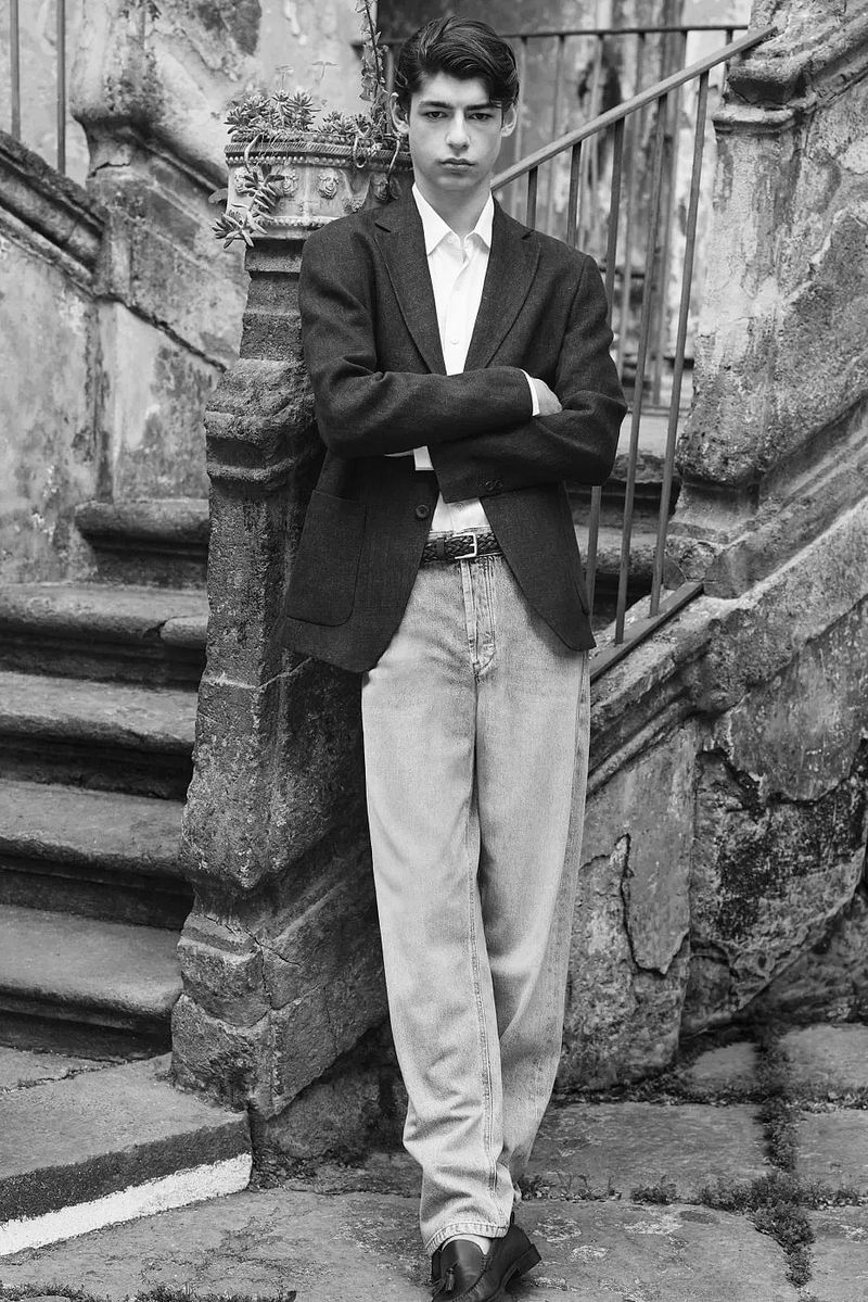 Man in a dark tailored blazer, white collared shirt, and relaxed light denim jeans standing by a weathered stone staircase.