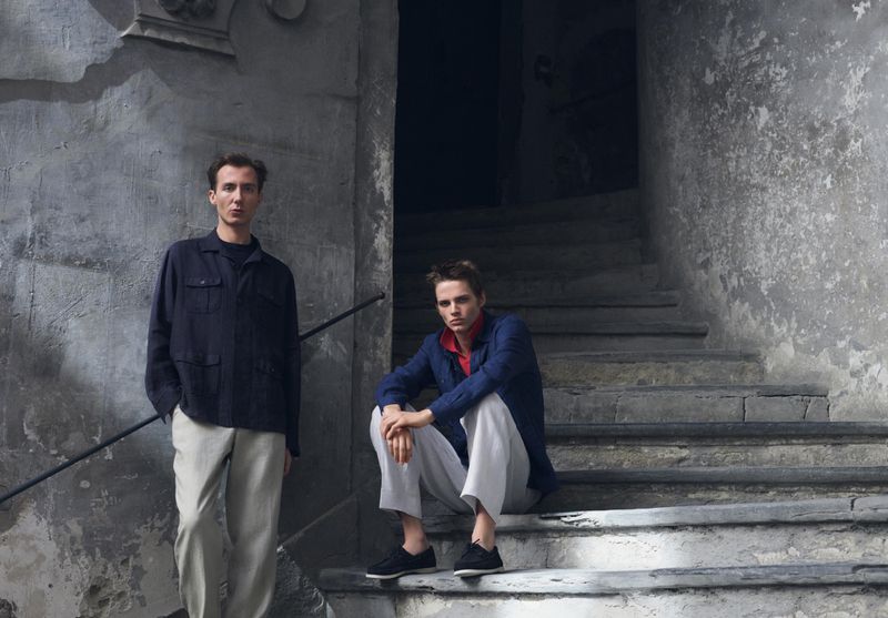 Two male models posing on weathered stone steps wearing relaxed buttoned overshirts and light trousers.