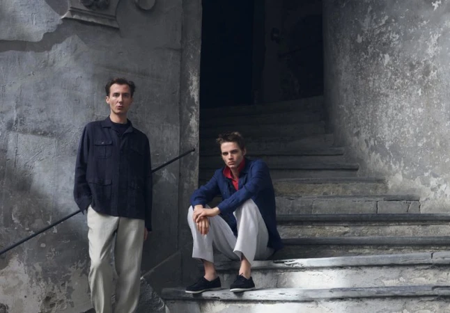 Two male models posing on weathered stone steps wearing relaxed buttoned overshirts and light trousers.