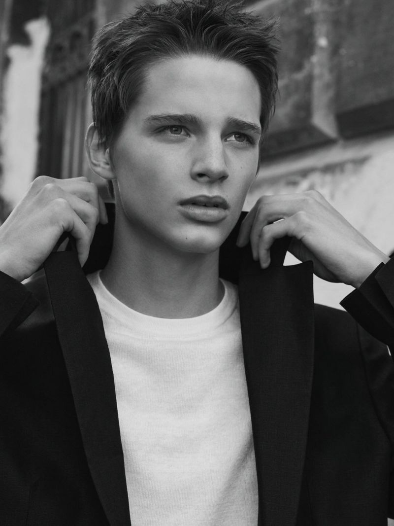 Black and white portrait of a young man adjusting the collar of his dark jacket over a white t-shirt outdoors.