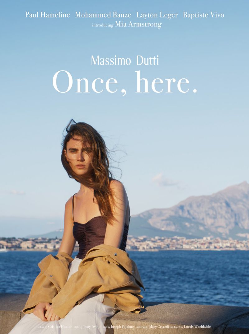 Model Mia Armstrong wearing a brown top, camel jacket, and white trousers seated against a coastal ocean backdrop for Massimo Dutti.