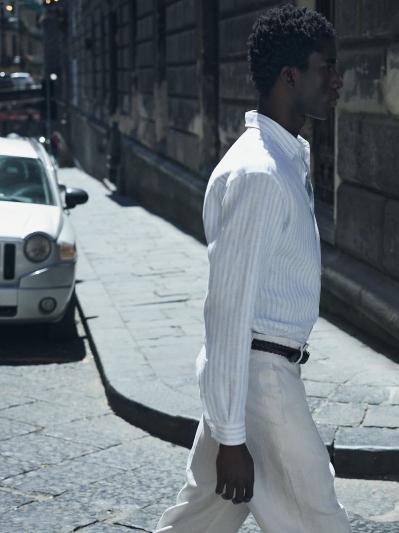 Profile view of a man walking down a sunlit city street wearing a light striped button-down shirt and relaxed white trousers.
