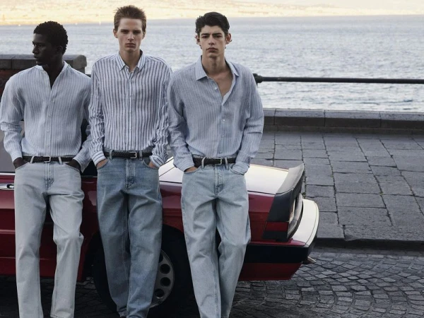 Three male models wearing striped shirts and light wash denim jeans leaning against a red car by the water.